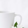 A stylish ceramic coffee cup for her, featuring an elegant floral design and a personalized name in a delicate script font, filled with steaming hot coffee, resting on a wooden coaster next to a small potted plant.