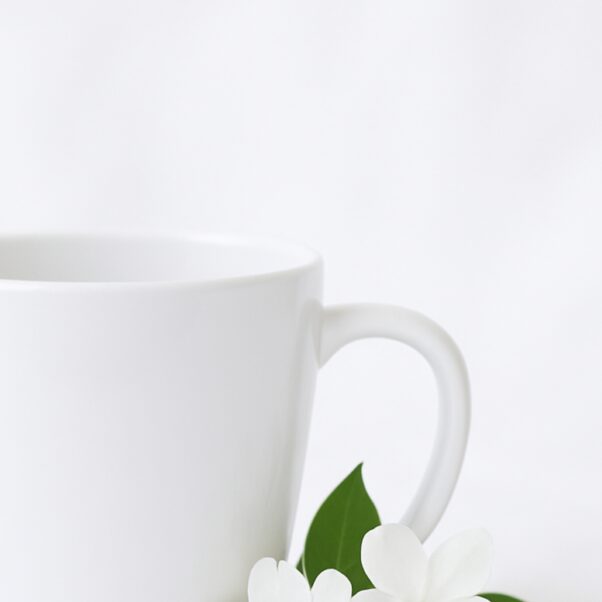 A stylish ceramic coffee cup for her, featuring an elegant floral design and a personalized name in a delicate script font, filled with steaming hot coffee, resting on a wooden coaster next to a small potted plant.