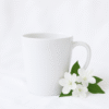 A stylish ceramic coffee cup for her, featuring an elegant floral design and a personalized name in a delicate script font, filled with steaming hot coffee, resting on a wooden coaster next to a small potted plant.