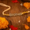 Brother Rakhi 4 A close-up of a "Brother Rakhi" on a wrist, featuring "BROTHER" spelled out in sparkling, studded gold-toned letters, flanked by decorative golden beads on a subtle, light-colored thread, symbolizing deep sibling affection.