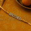 Brother Rakhi 2 A close-up of a "Brother Rakhi" on a wrist, featuring "BROTHER" spelled out in sparkling, studded gold-toned letters, flanked by decorative golden beads on a subtle, light-colored thread, symbolizing deep sibling affection.