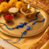 A "Bhaiya Bhabhi Evil Eye Rakhi Set" displayed on a golden plate, featuring one Rakhi with a simple blue evil eye bead for the brother and another (Lumba Rakhi) with a Hamsa hand charm and blue evil eye for the sister-in-law,