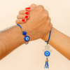 A "Bhaiya Bhabhi Evil Eye Rakhi Set" displayed on a golden plate, featuring one Rakhi with a simple blue evil eye bead for the brother and another (Lumba Rakhi) with a Hamsa hand charm and blue evil eye for the sister-in-law,