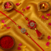 A "Traditional Floral Stone Bhaiya Bhabhi Rakhi Set" with a brother's Rakhi on a red thread featuring a round floral charm with red and white stones, and a matching Lumba Rakhi for the sister-in-law with a larger, ornate floral charm and red tassels, symbolizing love and prosperity.