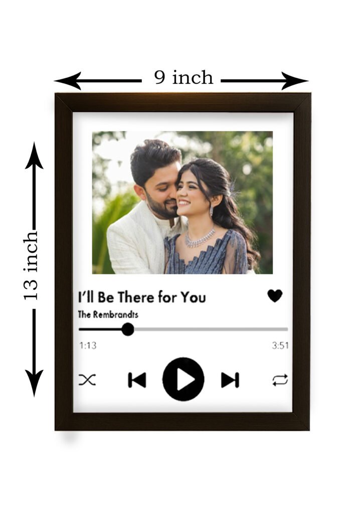 A framed Personalized Spotify Frame hanging on a white brick wall, featuring a custom photo of a couple, song title "I'll Be There For You," artist "The Rembrandts," and Spotify player controls, illuminated by a yellow hanging lamp.