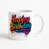 A white ceramic "Happy Birthday Mug" featuring a colorful, dripping paint-style "Happy Birthday" text, placed on a wooden desk with a blurred plant and papers in the background.