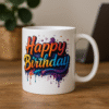 A white ceramic "Happy Birthday Mug" featuring a colorful, dripping paint-style "Happy Birthday" text, placed on a wooden desk with a blurred plant and papers in the background.