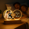 A glowing Personalized Wedding LED Lamp in the shape of two intertwined rings, featuring an etched photo of a wedding couple, text "Happy Wedding," and names "Saima & Naved," with "Forever in my heart" on the wooden base, set against a blurred background of a couple sleeping.