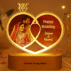 A glowing Personalized Wedding LED Lamp in the shape of two intertwined rings, featuring an etched photo of a wedding couple, text "Happy Wedding," and names "Saima & Naved," with "Forever in my heart" on the wooden base, set against a blurred background of a couple sleeping.