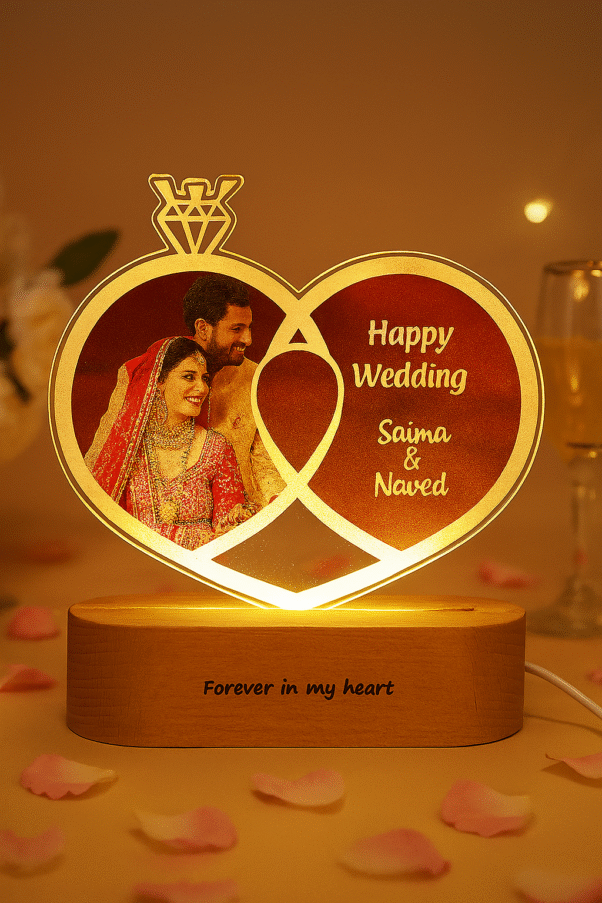 A glowing Personalized Wedding LED Lamp in the shape of two intertwined rings, featuring an etched photo of a wedding couple, text "Happy Wedding," and names "Saima & Naved," with "Forever in my heart" on the wooden base, set against a blurred background of a couple sleeping.
