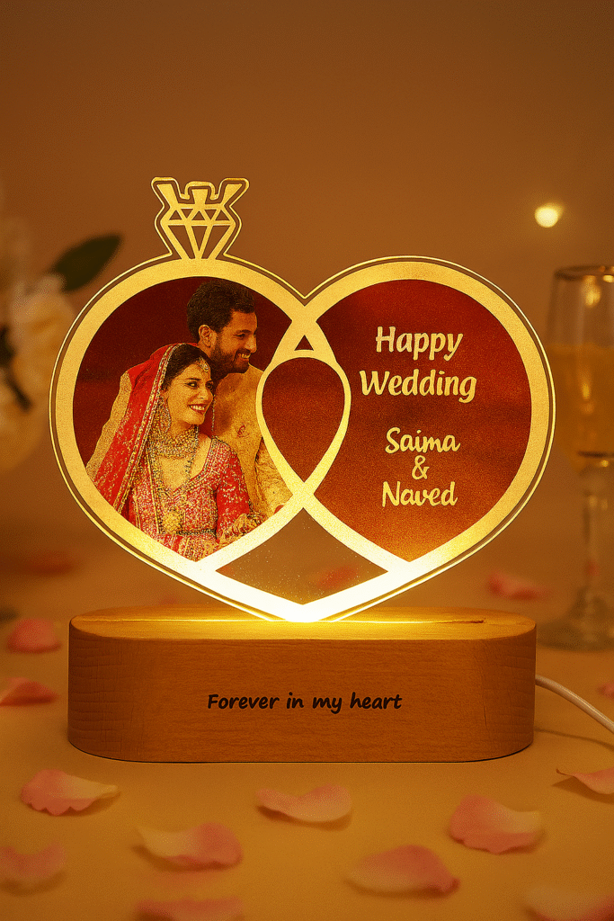 A glowing Personalized Wedding LED Lamp in the shape of two intertwined rings, featuring an etched photo of a wedding couple, text "Happy Wedding," and names "Saima & Naved," with "Forever in my heart" on the wooden base, set against a blurred background of a couple sleeping.