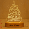 A glowing Personalized Birthday Acrylic LED Lamp shaped like a multi-tiered birthday cake with a candle on top, text "HAPPY BIRTHDAY," and a customizable "Add Name" section on its wooden base, set against a warm, blurred background.