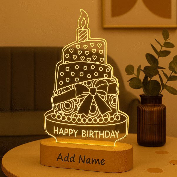 A glowing Personalized Birthday Acrylic LED Lamp shaped like a multi-tiered birthday cake with a candle on top, text "HAPPY BIRTHDAY," and a customizable "Add Name" section on its wooden base, set against a warm, blurred background.