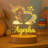 A glowing Customized Unicorn Lamp with Name on a wooden base, featuring an etched illustration of a unicorn with a rainbow mane and tail, surrounded by stars, with the name "Ayesha" personalized at the bottom.
