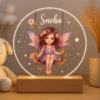A glowing circular Customized Acrylic LED Lamp with Name on a wooden base, featuring an etched illustration of a fairy on a swing surrounded by stars and flowers, with the name "Sneha" personalized at the top.