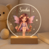 A glowing circular Customized Acrylic LED Lamp with Name on a wooden base, featuring an etched illustration of a fairy on a swing surrounded by stars and flowers, with the name "Sneha" personalized at the top.