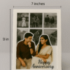 A white-framed Romantic Anniversary Photo Collage Frame featuring a main photo of a couple and smaller surrounding photos, with "Happy Anniversary" text, on a wooden table next to a vase
