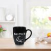 A black Bro No 1 Best Brother Mug featuring text and a white bicep icon, placed on a wooden counter with white ceramic rabbit decorations and flowers in the background.