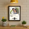A framed Personalized Spotify Frame hanging on a white brick wall, featuring a custom photo of a couple, song title "I'll Be There For You," artist "The Rembrandts," and Spotify player controls, illuminated by a yellow hanging lamp