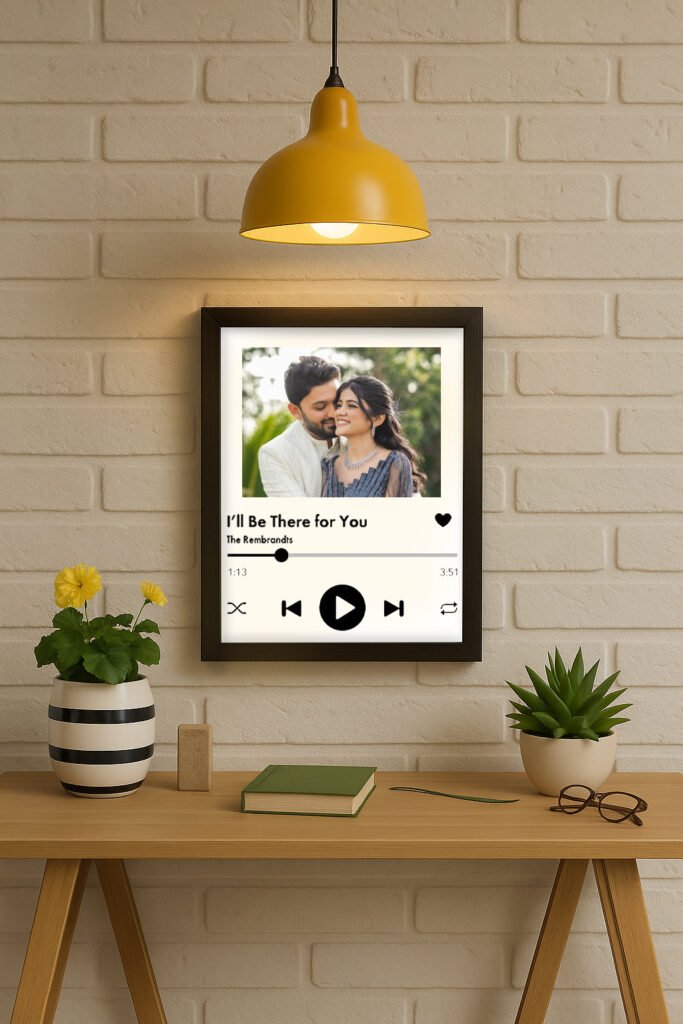 A framed Personalized Spotify Frame hanging on a white brick wall, featuring a custom photo of a couple, song title "I'll Be There For You," artist "The Rembrandts," and Spotify player controls, illuminated by a yellow hanging lamp