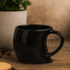 Black singapore mug with a custom 'CafeWorld' logo in orange and white, placed on a wooden desk next to a laptop and a notebook.