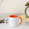 Orange and White Ceramic Coffee Mug a bright orange interior and handle, sitting on a table with an antique clock.
